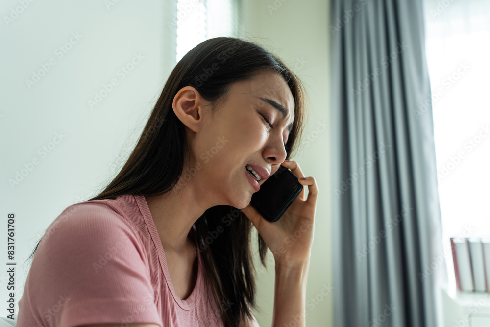 Wall mural asian upset depressed woman sitting on bed and talking on mobile phone.