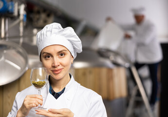 Portrait of positive woman brewmaster holding glass of hops pellets in beer factory.