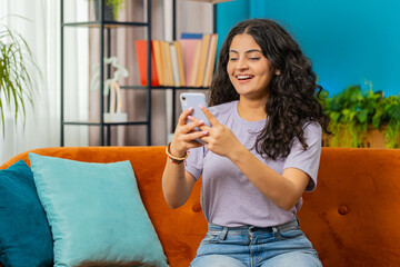 Indian woman sits on couch uses mobile phone smiles at home living room apartment. Young Arabian...