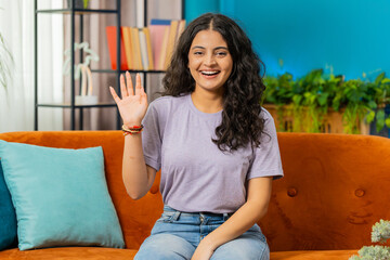 Hello. Indian pretty woman smiling friendly at camera and waving hands gesturing hello hi, greeting or goodbye welcoming with hospitable expression at home. Arabian girl sitting on sofa in living room
