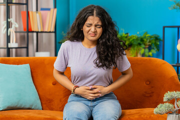 Sick ill Indian woman suffering from period cramps, painful stomach ache on sofa at home room. Girl...