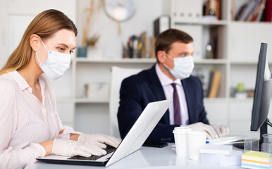 Focused young woman in medical face mask and latex gloves working on laptop in office with male...