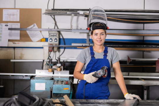 Confident smiling young woman in blue overalls, protective face shield and gloves standing with handheld angle grinder at profile cutting table in window production and assembly workshop.. - Powered by Adobe