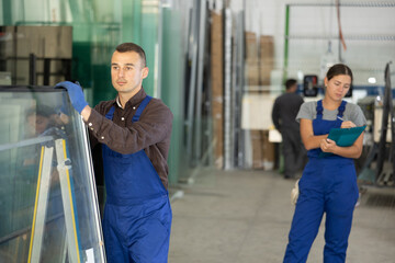 Focused skilled male worker in blue overalls moving insulated glass units on rack across factory...
