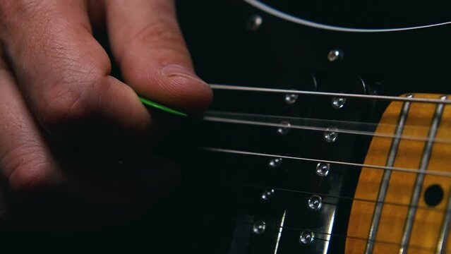 Close-up of a man's fingers with a green plectrum striking the top two strings of an electric guitar. The guitar has a black body. Slow motion of the vibration of the strings