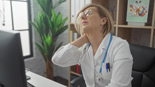 A mature, caucasian, blonde, female doctor in a clinic room, experiencing neck pain while seated at her workstation.