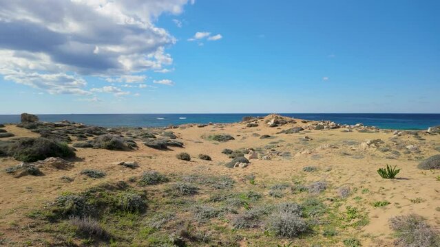 A wide view of the sandy terrain at the Tombs of the Kings in Pafos, Cyprus, with the sea in the background and a partly cloudy sky