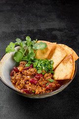 Chilli con carne with beans and corn tortilla on black background. Traditional mexican food - chilli with minced meat and spices in bowl on dark backdrop. Chili con carne in minimal style.