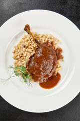 Classic dish french cuisine - roasted duck leg in porto sauce with garnish on black background. Cooked duck leg with pearl barley on white plate on dark concrete table. Duck confit in minimal style.