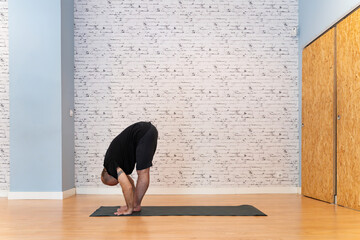 Senior man in black outfit practicing a deep forward bend yoga pose on a mat in a bright, spacious room with a white brick wall and wooden floor. 