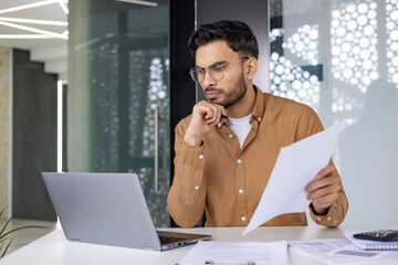 Focused businessman working on laptop and holding documents in modern office