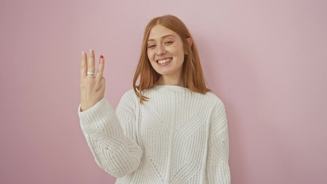 Joyful young redhead woman confidently pointing upward, showing number three with fingers all while positively beaming. captured against an isolated pink background.