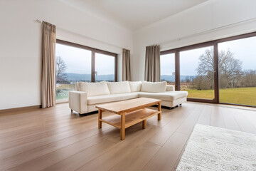 Stylish, bright, open living room interior with large windows and lots of natural light. White corner sofa,  solid oak wood coffee table and a white rug.