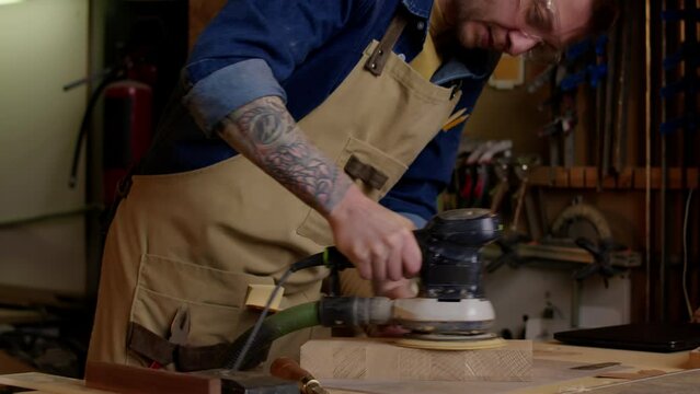 Medium shot of Caucasian male carpenter wearing safety glasses and earmuffs using angle grinder on wooden piece while working in workshop
