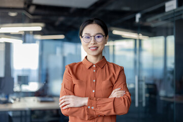 Confident businesswoman with glasses standing with arms crossed in a modern office setting,...