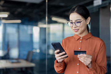 A businesswoman in a modern office conducts an online payment using her smartphone and credit card, illustrating technology and finance.