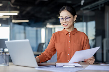 Young professional woman working on a laptop in a modern office setting, holding documents and focused on her tasks.