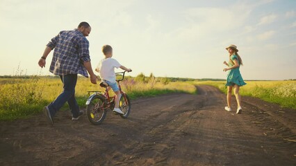 Childhood bike concept. Mother and dad teaching son to ride bicycle. Young family teaching son to ride bike first time on countryside rural road at sunset. Happy kid concept. The best moments together