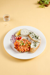 Salmon and avocado tartare with crispy bread and cream cheese, elegantly plated with lemon and dill on a white plate against a yellow background