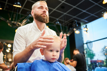 Young child getting haircut in modern barbershop. Hairdresser making final touches in new haircut,...