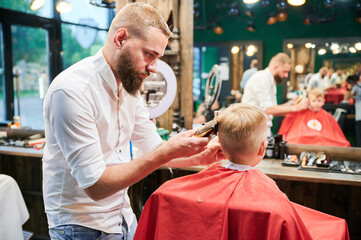 Barber using comb and shaver to cut hair. Professional hairdresser shaving little kid's nape.