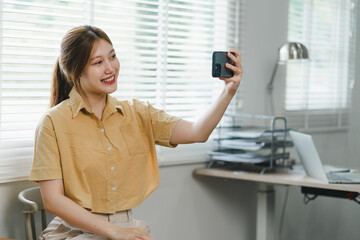 Young woman in yellow shirt smiles brightly while taking a selfie in her home office.