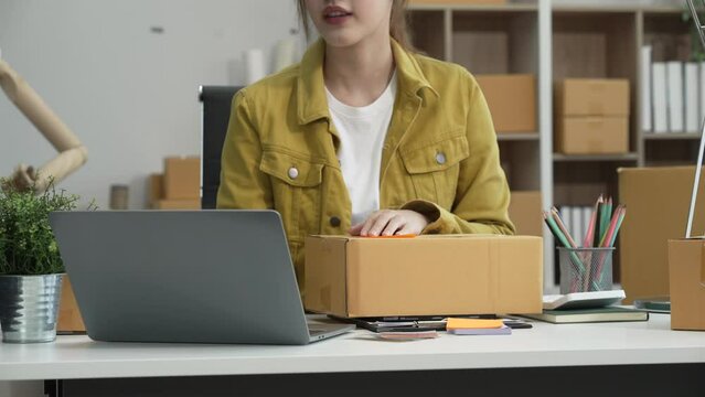 A young Asian businesswoman efficiently manages SMEs, utilizing her desk, tablet, laptop to handle online orders and live streams, showcasing entrepreneurial prowess in the dynamic market landscape.