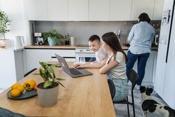Asian teenage girl spending time with her little brother sitting in the kitchen and teaching how to use a laptop