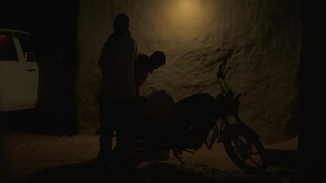 Two african men remove large tank from motorcycle near wall of house late in evening. Hard daily life in slums