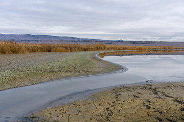 The curved river bed flows into the lake. Around cracked earth and tall grass. Reflections of sky