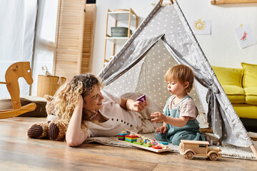 Curly mother and toddler daughter happily playing together inside a colorful play tent, embracing...