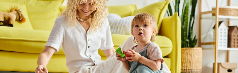 Naklejka premium A curly mother and her toddler daughter stand beside a yellow couch at home, following the Montessori method of education.