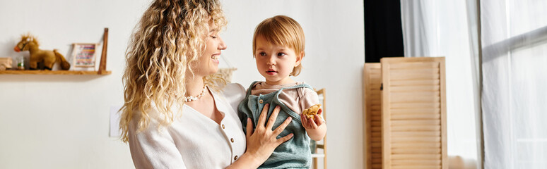 A curly mother and her toddler daughter bask in the morning sunlight, standing together in front of a window.