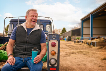 Mature Male Farm Worker Sitting On Tailgate Of Off Road Farm Vehicle Holding Hot Drink In Cup