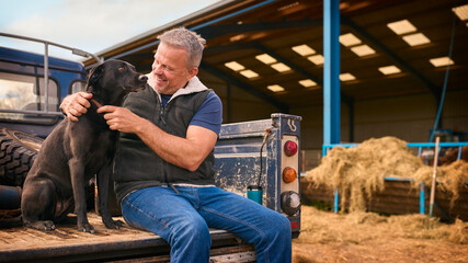 Mature Male Farm Worker Sitting On Tailgate Of Off Road Farm Vehicle With Dog