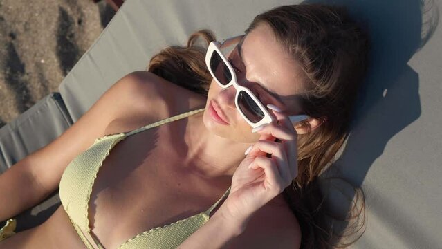 Beautiful woman in sunglasses resting on sunbed at beach, above view. Camera moving around girl