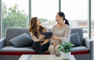 Two women are sitting on a couch, laughing together and sharing a tablet computer, in a bright...