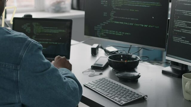 Back view slowmo of Black male system administrator sitting in front of laptop and two computer monitors with html codes on black screen while working in office