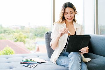 Young Asian woman designer sitting and using sample color swatches and digital tablet with pencil to sketch and design the new project. Fashion designer working in studio. Small business concept.