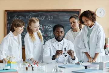 Young African American man wearing lab coat sitting at desk in classroom teaching group of kids how to use microscope in Chemistry class