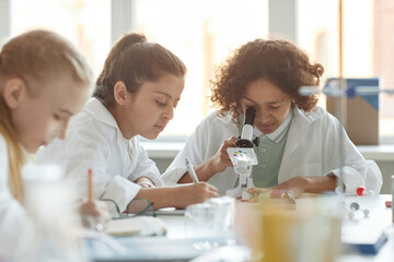 Selective focus shot of ethnically diverse middle school students wearing lab coats using microscope and making note during Chemistry lesson