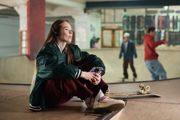 Modern Caucasian girl sitting on floor in skatepark holding retro portable audio player enjoying listening to music, copy space