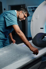 Mature biracial male radiographer preparing CT scanner bed for next patient at work in modern...