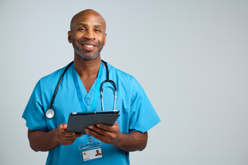 Studio Portrait Shot Of Smiling Mature Doctor Wearing Scrubs Using Digital Tablet © Monkey Business