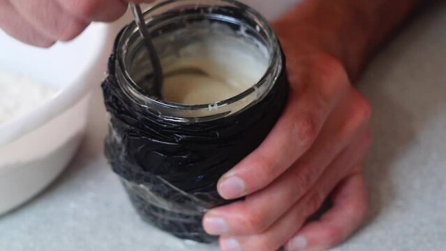 Kneading liquid yeast-free starter in a glass jar shaded with a black bag by hand with a metal tablespoon by the hands of a white man. The concept of baking according to an old recipe.