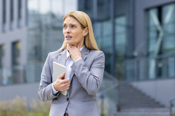 Fototapeta premium Businesswoman in a gray suit clutching her throat in discomfort while holding a tablet outside an office building.