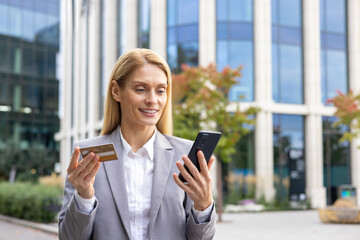 Smiling businesswoman using a credit card and smartphone for online payment. Outdoor shot with modern building in the background.