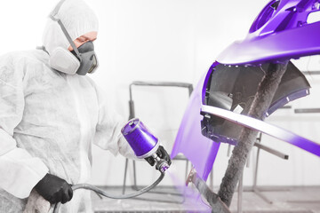Auto mechanic worker painting car bumper with spray gun in a paint chamber during repair work.