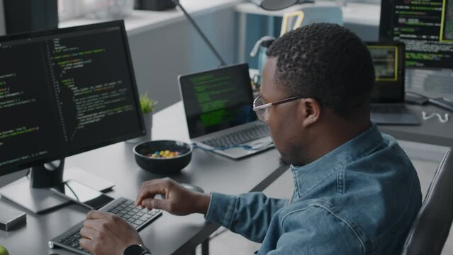 Chest up of young Black man in eyeglasses and denim shirt program coding on desktop computer while working in modern IT office