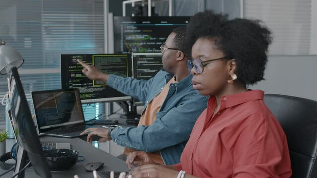 Medium shot of young African American couple of software engineers sitting at desk in font of computer monitor and having discussion, working together in modern office, coding and developing programs